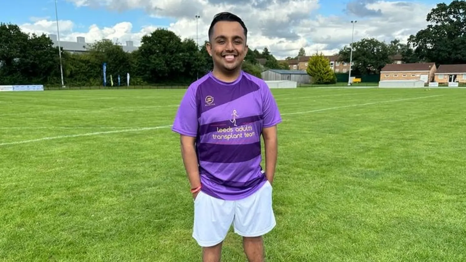 A young Asian man in a t-shirt, with an alternating light/dark purple pattern, stands in the middle of a sports field. His t-shirt says "Leeds adults transplant team" in gold letters. He's smiling with his hands in his white shorts pockets. The field around him looks immaculately well-kept, with white lines painted on it. In the distance a small, beige clubhouse building is visible.