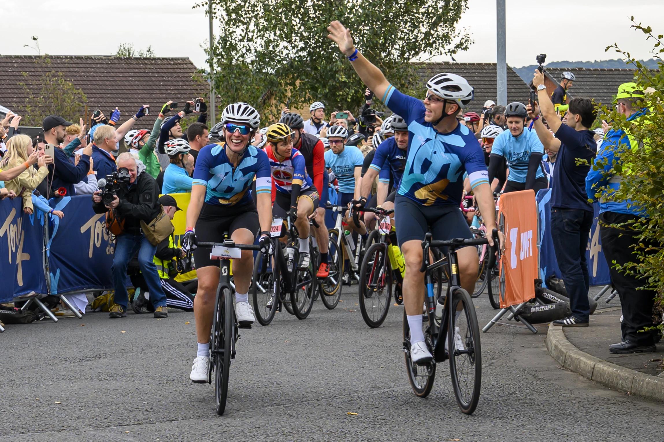 Sir Chris Hoy and wife Sarra wave to onlookers as they ride bikes down a road.