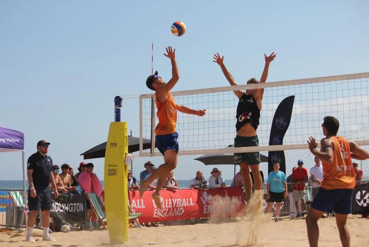A beach volleyball match taking place on a sunny day. Two players - one in an orange vest and one in a dark vest - are both jumping at the net, reaching up toward a volleyball. Another player in an orange vest is standing nearby. Spectators are watching from behind a barrier, with beach umbrellas, banners and flags visible in the background. The sea and clear blue sky are also visible behind the court.