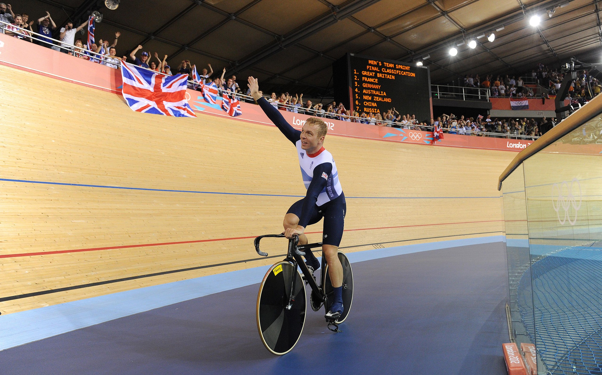 Sir Chris Hoy waves while riding a bike in a velodrome