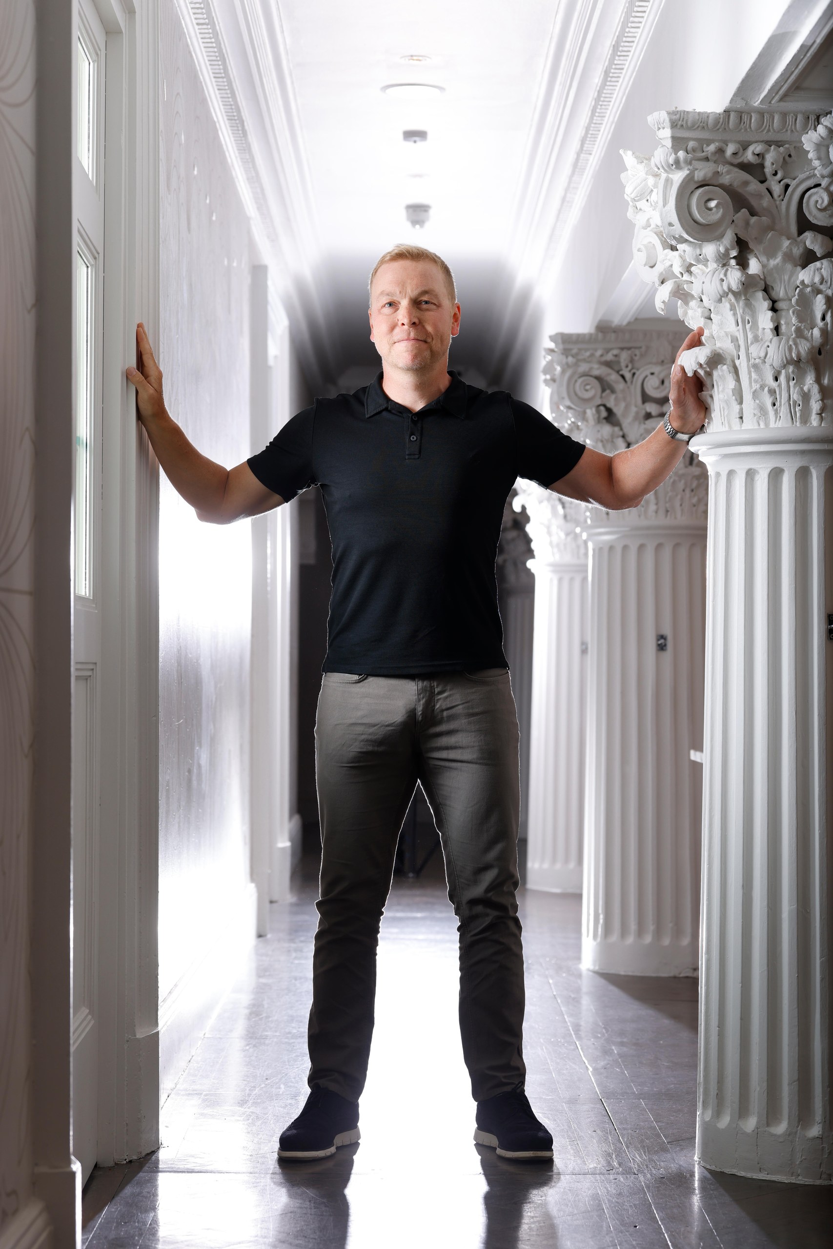Sir Chris Hoy stands between a pillar and a wall in a corridor with his arms outstretched.